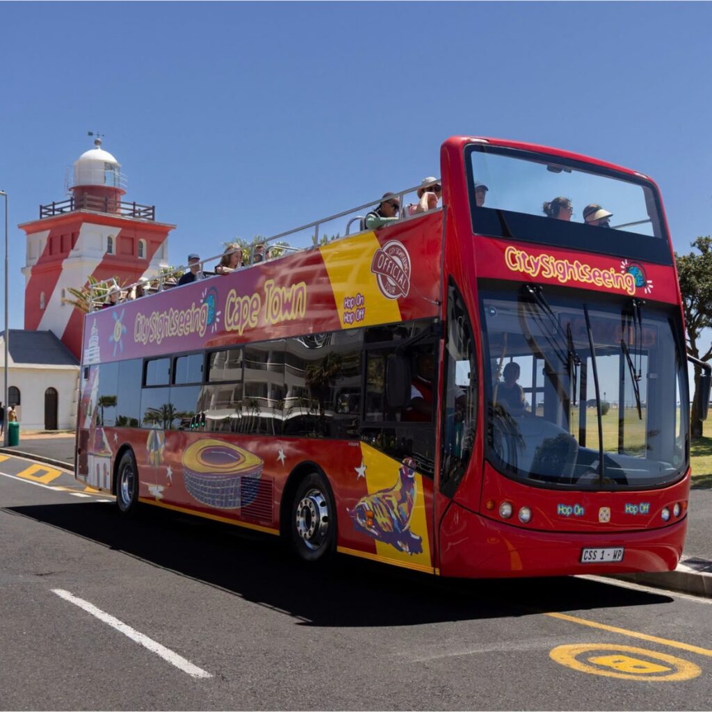 Red City Sightseeing open-top bus beside a red-and-white lighthouse in Cape Town