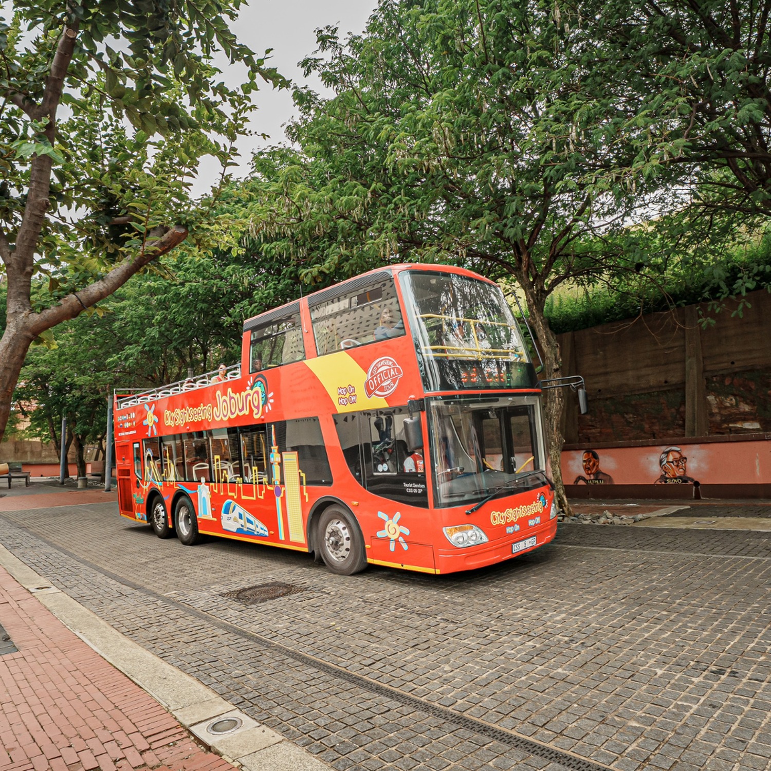 Red City Sightseeing Johannesburg double-decker bus parked on a tree-lined cobbled street
