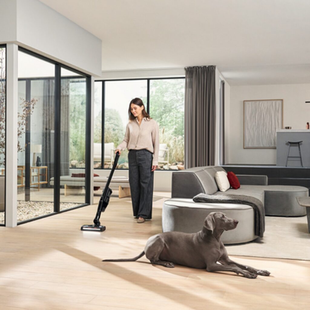 Woman using a Miele cordless vacuum in a bright living room with a dog nearby